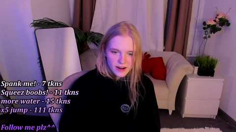 Media: Video of a young, fair-skinned woman with long blonde hair, wearing a black turtleneck, seated in a modern living room with a white couch, potted plants, and purple curtains. Text overlay reads: \"Spam me: 7 7 7 7 7 7 7 7 7 7 7 7 7 7 7 7 7 7 7 7 7 7 7 7 7 7 7 7 7 7 7 7 7 7 7 7 7 7 7 7 7 7 7 7 7 7 7 7 7 7 7 7 7 7 7 7 7 7 7 7 7 7 7 7 7 7 7 7 7 7 7 7 7 7 7 7 7 7 7 7 7 7 7 7 7 7 7 7 7 7 7 7 7 7 7 7 7 7 7 7 7 7 7 7 7 7