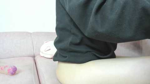 Video of a person in black sweatpants, sitting on a pink couch. The background is a plain white wall, and a white stuffed animal rests on the couch.