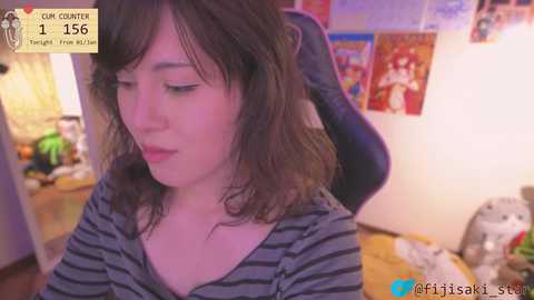 Video of a young woman with shoulder-length brown hair, wearing a striped shirt, sitting at a desk with posters and toys in a cluttered room.