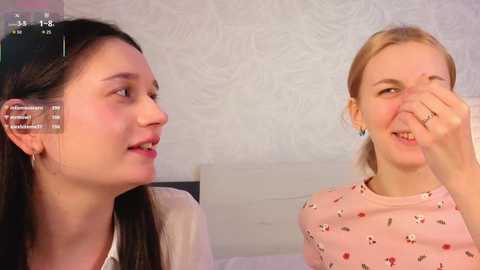 Video of two young women with fair skin, one with long dark hair and another with blonde hair. They are laughing, sitting in a bedroom with a white, textured wallpaper background.
