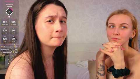 Video of two young women with long hair: one with black hair, the other with blonde. Both appear distressed, holding their hands together. Background shows a bed with white sheets.