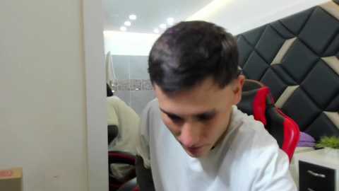 Video of a young man with short dark hair, wearing a white T-shirt, sitting on a black and red gaming chair in a modern room with a tiled bathroom and LED lights.