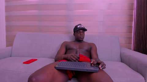 Video of a shirtless, muscular Black man with a cap, red shorts, and a laptop, sitting on a light-colored couch against a striped wall.