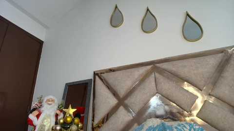 Video of a cozy bedroom corner featuring a plush, beige upholstered headboard with a diamond pattern. Three decorative, gold-framed, tear-drop mirrors adorn the wall above. To the left, a Santa figurine stands beside a festive, colorful Christmas tree with golden ornaments.