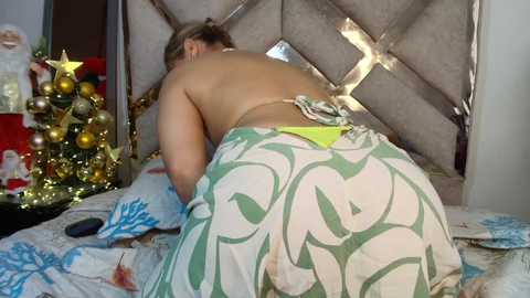 Video of a topless woman with light skin, blonde hair tied back, wearing a white skirt with green floral pattern, leaning forward on a bed with a gold and silver Christmas tree and presents in the background.