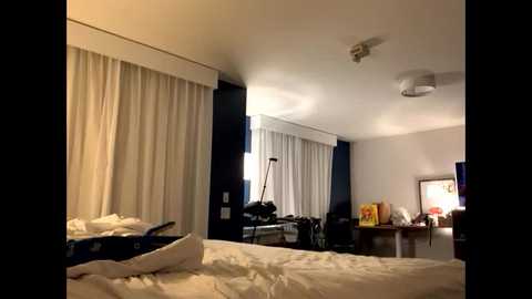 Video of a dimly lit hotel room with white curtains, a bed with rumpled sheets, a desk with a computer monitor, and a TV on a stand.