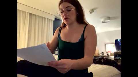 Video of a fair-skinned woman with glasses, long red hair, in a dark green tank top, reading a document in a dimly lit hotel room with beige curtains, a TV, and a bed.