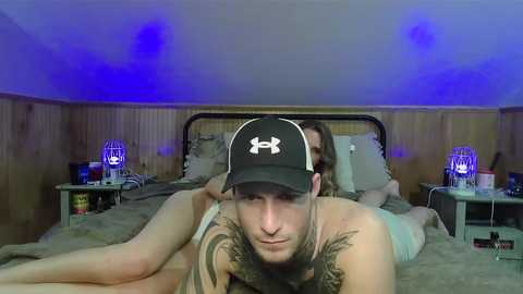 Video of a man with a beard and tattoos on his chest, wearing a black cap, lying on a bed with a wooden headboard, in a dimly lit room with blue lights.
