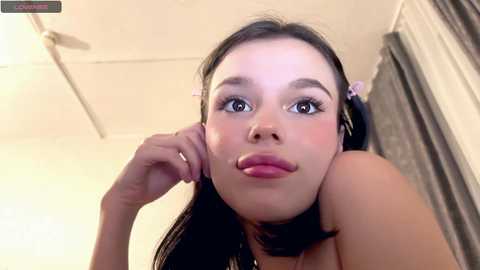 A young woman with light skin, long black hair in pigtails, wearing a pink headband, makes a playful face. She has pink lipstick and is wearing headphones.