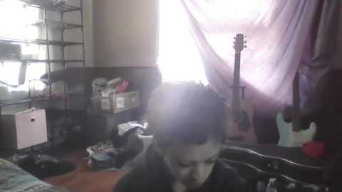 A video shows a boy with dark hair in a messy room with a guitar, pink fabric, and a cluttered bookshelf.