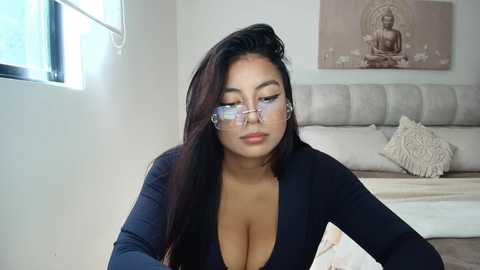 Media: Video of a young Asian woman with long black hair, wearing glasses and a low-cut dark blue top, sitting on a bed in a modern bedroom with a Buddha wall art.