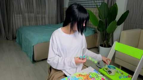 Video of a woman with black hair, wearing a white long-sleeve shirt, painting on a table in a modern, light-colored bedroom.