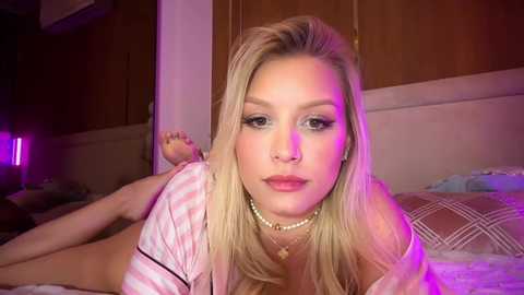 Video of a blonde woman with fair skin, wearing a pink striped top, lying on a bed with a pink and white geometric-patterned pillow. Background includes wooden cabinets and a dimly lit room with purple lighting.