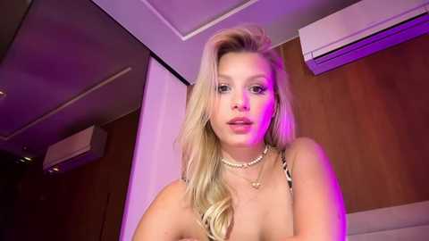 Video of a blonde woman with fair skin and long hair, wearing a black and white striped top and pearl necklace, in a dimly lit room with purple lighting, air conditioning unit on the wall.