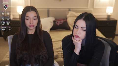 Video of two women, one with long, straight, dark hair, and the other with long, wavy, dark hair, seated in a modern bedroom with beige walls, a bed, and lamps.