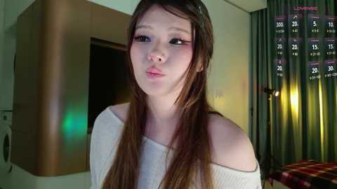 Video of a young Asian woman with long brown hair, wearing a white off-shoulder top, standing in a modern kitchen with green curtains, a calendar, and a red and yellow checkered towel in the background.