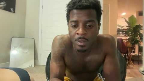 Video of a shirtless, muscular Black man with short hair and a mustache, wearing yellow shorts, sitting on a carpet in a dimly lit room with a mirror, potted plant, and doorway.