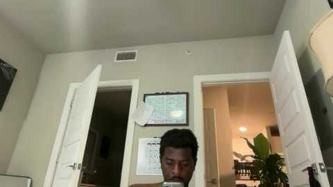 Video of a man with short black hair, standing in a small, dimly lit room with beige walls. He is looking down, holding a white piece of paper. The room has a table, a door, and a framed calendar.