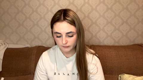 Media: A video of a young woman with light skin and long brown hair, wearing a white sweatshirt, sitting on a brown couch in a room with a beige patterned wallpaper.