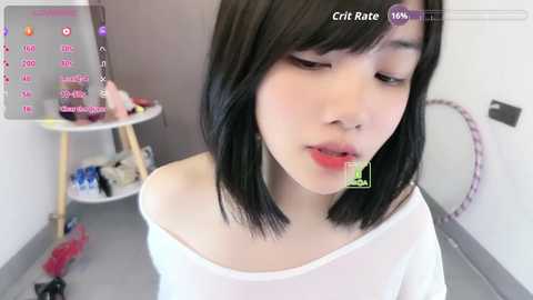 Media: A video of a young Asian woman with straight black hair, wearing a white off-shoulder top, standing in a minimalistic room with a white wall and a small table.