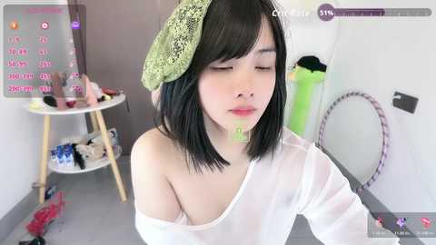 Media: A video of a young Asian woman with black hair, wearing a white off-shoulder shirt, sitting in a messy room with a green lace headband and food on her face.