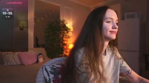 Video of a young woman with long brown hair, seated indoors, wearing a floral shirt. The background features a cozy living room with a TV, a potted plant, and a warm, ambient light.