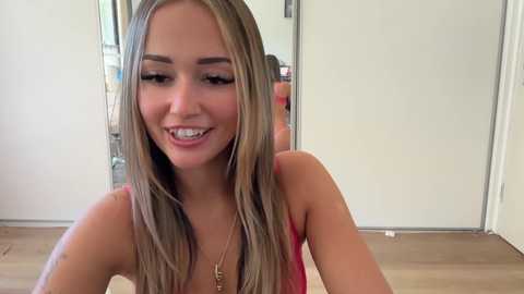 Media: Video of a smiling, slender young woman with straight, blonde hair, wearing a red tank top, in a bright, modern room with wooden floors and white walls.
