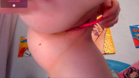 Video of a fair-skinned, nude person with medium-sized breasts, pulling a pink thong aside, in a room with colorful, comic book-style posters and a blue ball on a bed.