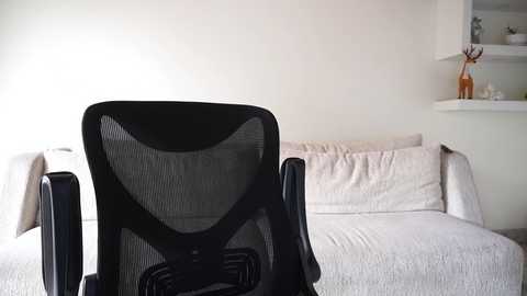A video of a modern, minimalist living room featuring a black mesh office chair facing a beige sectional sofa with light-colored pillows and a white wall.
