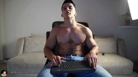 Video of a shirtless, muscular Asian man with short black hair, wearing jeans, seated in a beige sofa, using a keyboard.