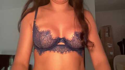 Video of a woman with long brown hair wearing a lacy, blue bra with intricate floral patterns, revealing medium-sized breasts. The background shows a dimly lit, indoor setting with a white wall and some indistinct furniture.