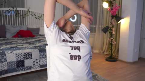 Video of a woman stretching in a cozy bedroom with a white bed, floral curtains, and a floor lamp.