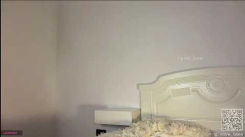 A video of a minimalist, cream-colored bedroom with a decorative headboard, a white wall, and a fluffy white bedspread.