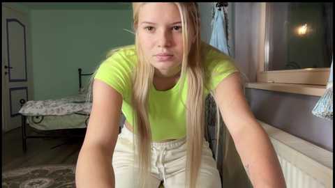 Video of a young blonde woman with fair skin wearing a lime green t-shirt and white pants, leaning forward in a dimly lit bedroom.