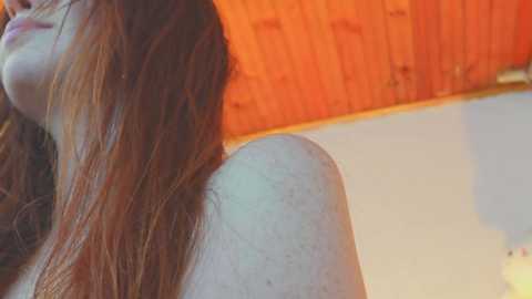 A close-up video of a young woman with long, auburn hair, partially visible, in a bright room with warm, orange wooden walls and a white floor.