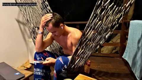 Video of a shirtless man with short dark hair, wearing blue shorts and headphones, sitting on a wooden floor, looking at a laptop.