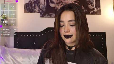 Video of a young woman with long brown hair, wearing black makeup, a black choker, and a black top, sitting on a bed with a black headboard and purple bedding, surrounded by dark artwork.