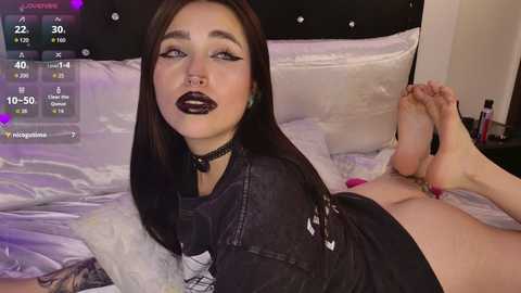 Video of a pale-skinned woman with long dark hair, black lipstick, and dark eye makeup, lying on a bed with white sheets, wearing a black choker and a black shirt, with a digital thermometer screen overlay.