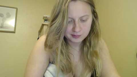 Video of a young Caucasian woman with long, wavy blonde hair, wearing a white bra, sitting in a room with beige walls, a framed picture, and a bookshelf.