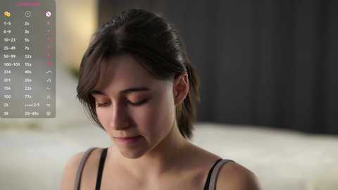 Video of a young Asian woman with short, dark hair, wearing a black spaghetti-strap top, sitting with eyes closed in a dimly lit room.