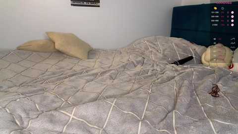 A video of a messy, unmade bed with a beige quilted comforter, two beige pillows, a pair of black glasses, and a black smartphone on the right side.