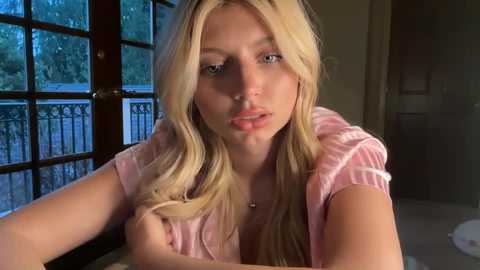 Video of a young blonde woman with long hair, wearing a pink top, leaning against a window with a view of greenery.