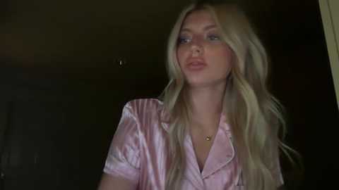 A video of a fair-skinned, blonde woman with long hair, wearing a pink satin shirt, standing indoors with dim lighting, looking slightly off-camera.
