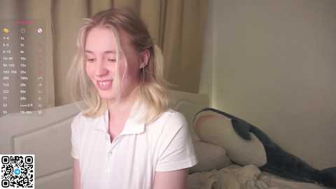 A video of a smiling young woman with blonde hair in a white shirt, lying on a bed with a plush shark toy beside her.