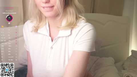 Video of a fair-skinned woman with blonde hair, wearing a white polo shirt, smiling slightly, sitting on a bed with white sheets, in a bedroom with a calendar and QR code visible.