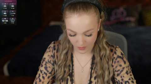 Video of a young woman with fair skin and wavy blonde hair, wearing a leopard-print top and black headband, sitting indoors, focused on a screen displaying game stats.