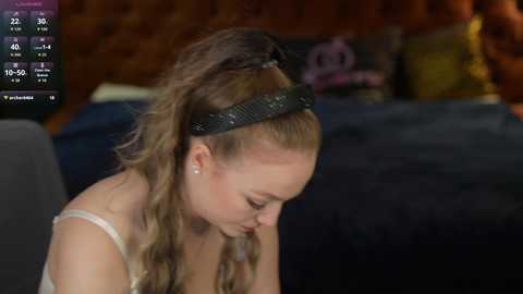 Video of a fair-skinned woman with long, wavy, blonde hair, wearing a black headband and a white spaghetti-strap top, sitting on a bed with dark bedding.