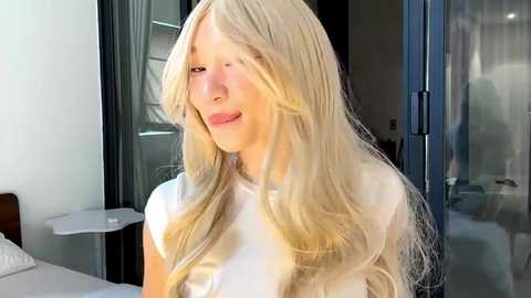 Video of a fair-skinned woman with long, wavy blonde hair, wearing a white top, smiling softly, standing in a modern, brightly lit room with large windows and glass doors.
