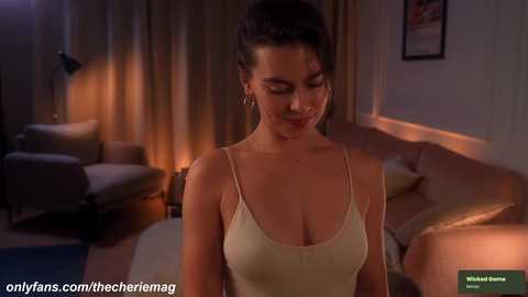 A video of a woman with medium skin tone, dark hair in a bun, wearing a beige camisole, standing in a dimly lit living room with beige curtains, a couch, and a lamp.