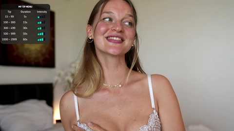 Video of a smiling Caucasian woman with light skin and long, straight blonde hair, wearing a lacy, white bra, in a bedroom with a bed and abstract wall art.